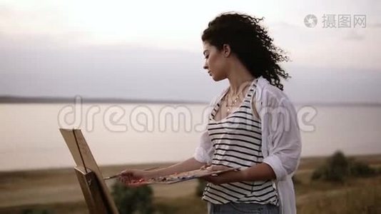 一位迷人的卷曲女艺术家在湖边的户外画架上画着她的画。 早晨，柔软视频