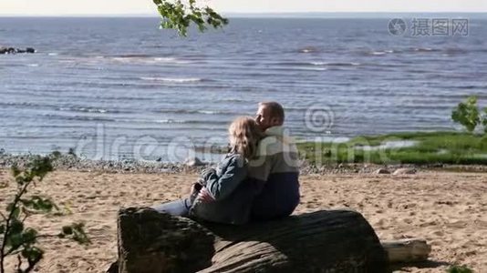 一位孕妇正在湖边拥抱她的丈夫，坐在原木上。 年轻家庭视频
