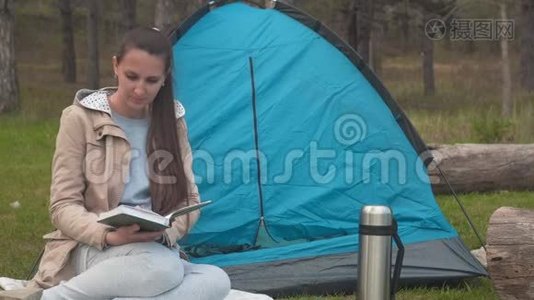 一个留着长发的小女孩正坐在森林里的一个蓝色帐篷附近看书..视频