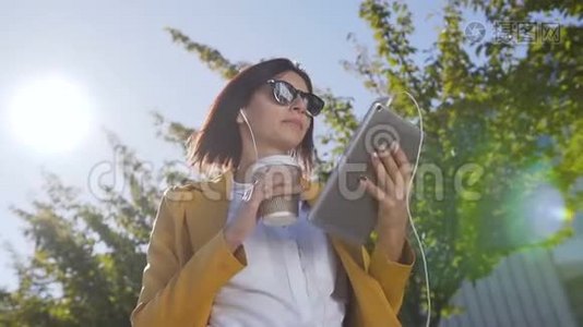 美丽的年轻女子戴着太阳镜，拿着热咖啡在杯子里，同时她在平板电脑上发送信息。 户外活动视频
