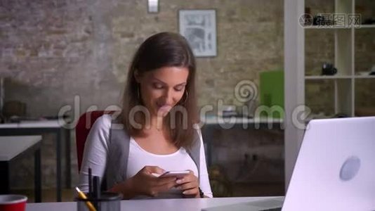 漂亮的女办公室工作人员坐在笔记本电脑前，在电话上发短信，笑了起来视频