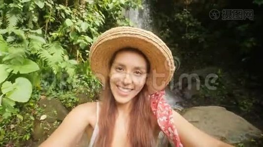 年轻的快乐混合种族旅游博客女孩穿着白衣服和草帽制作自拍视频使用手机在惊人的野生视频
