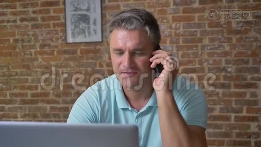 精力充沛的白种人成年男性在办公室里坐着用电话和电脑说话，棕色的砖块视频