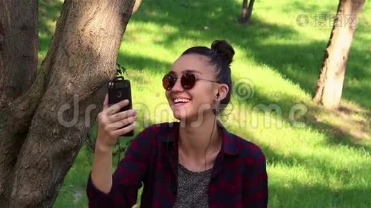 戴着太阳镜的漂亮女孩，积极地笑着，透过社交网络上的新闻，坐在公园的长凳上。视频