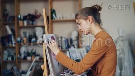 严肃的年轻女士艺术学生正在画室的画布上用画笔和颜料画花朵。 生活方式视频
