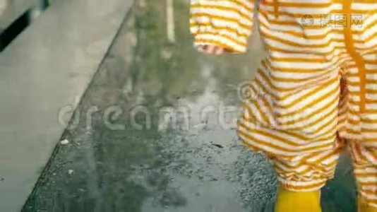 小宝宝穿着橙色条纹防水雨衣和黄色橡胶雨靴在雨后的水坑上散步，特写镜头视频