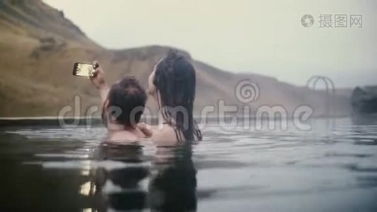 年轻夫妇在山区山谷的智能手机上拍照。 一男一女在冰岛温泉游泳..视频