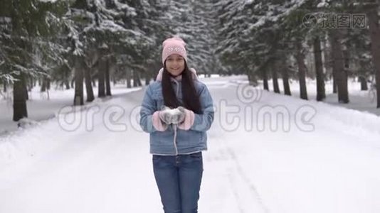 心情好的漂亮女孩站在冬天的森林里微笑着，用手吹雪。 慢速高清视频