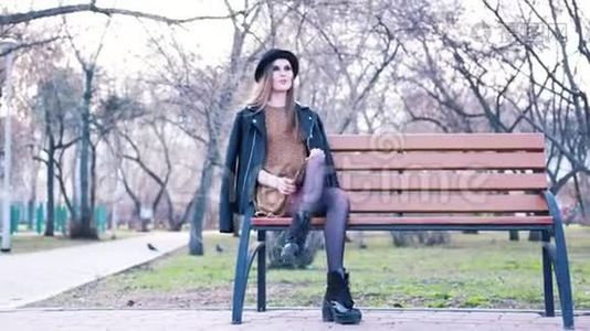 漂亮的年轻女子，穿着时尚服装，黑帽子，脚踝靴和皮夹克，独自坐在公园的长凳上。视频