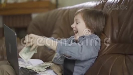 六岁的小男孩坐在皮沙发上，腿上放着笔记本电脑，把钱扔到空中视频