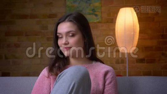一位年轻美丽的白种人白种人女性在舒适的沙发上坐着，看着摄像机，微笑着拍摄特写镜头视频
