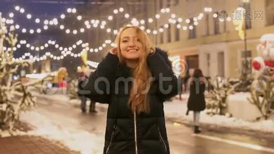 穿着冬季夹克的漂亮女孩站在冬季街道上闪烁的花环的背景上摩擦着她视频