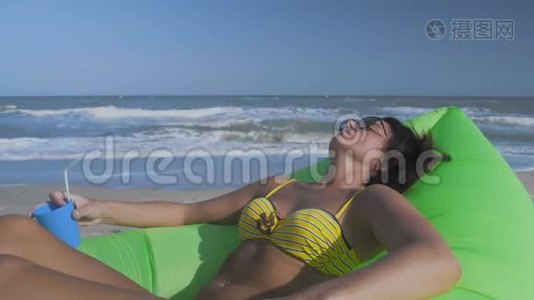 近景美丽的女人在比基尼日光浴海滩谎言绿色充气枕头与鸡尾酒在手视频