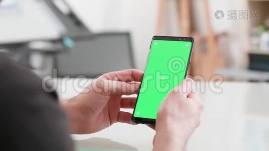 男人坐在办公桌前，手里拿着绿色屏幕的智能手机视频