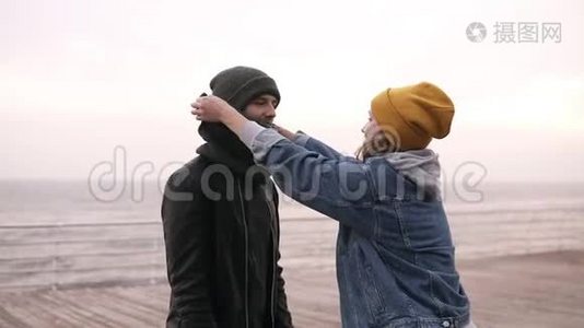 年轻可爱的一对夫妇，穿着温暖的衣服和帽子，一边站在海边一边，一边有趣地纠正他们的帽子视频