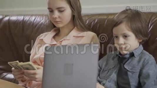 姐姐和弟弟坐在皮革沙发上。 那个男孩拿着笔记本电脑，女孩在数钱视频