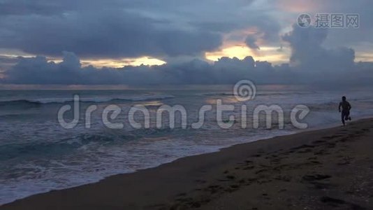 日落时，一个带着狗的年轻人沿着热带海洋的海滩奔跑的剪影视频