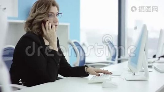 年轻的休闲女士坐在办公桌前，用笔记本电脑，接电话，打电话，微笑着在手机上说话视频
