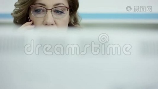 年轻的休闲女士坐在办公桌前，用笔记本电脑，接电话，打电话，微笑着在手机上说话视频