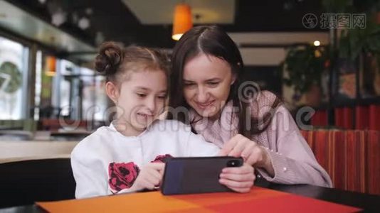 漂亮的白种人母亲和她的女儿在咖啡馆里使用智能手机。 一个先进的小女孩展示并解释视频