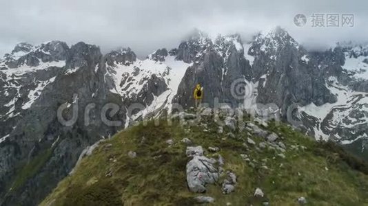 一个徒步旅行者站在山顶上，看着白雪皑皑的山脉，举起双手做胜利的手势。视频