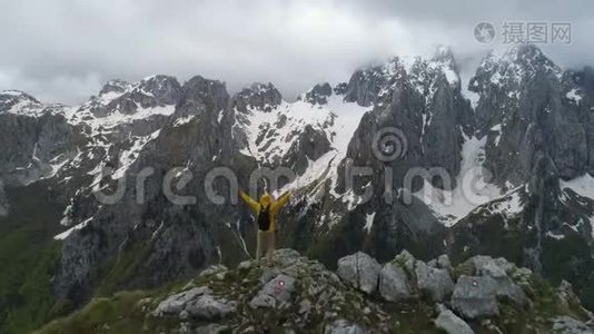 一个徒步旅行者站在山顶上，看着白雪皑皑的山脉，举起双手做胜利的手势。视频