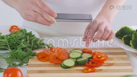 女人`手拿着智能手机，正在为沙拉拍摄新鲜蔬菜的照片，切片在切割板上。视频