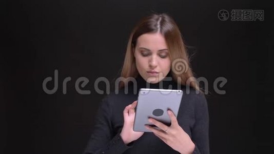年轻的白种人长发金发的金发女孩在黑色背景下专注地观看平板电脑的肖像。视频