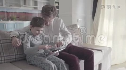 父亲和孩子坐在大客厅的沙发上，在平板电脑屏幕上滚动。 爸爸教他的孩子。 父子关系视频