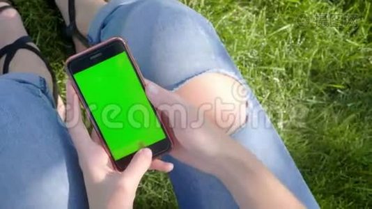 年轻漂亮的女孩穿着公园里的街服，撕破牛仔裤。 他手里拿着一部带有绿色屏幕的智能手机视频