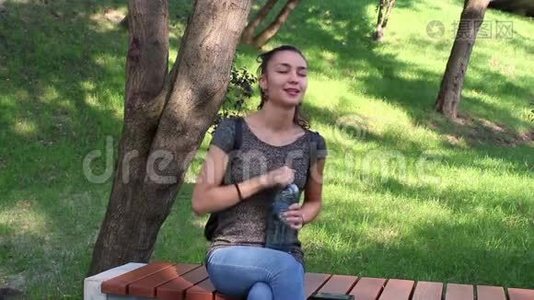 年轻漂亮的女孩用水摇动一个塑料瓶，坐在树荫下的长凳上。视频