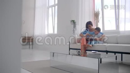 一位年轻的母亲带着一个孩子读一本书，坐在客厅里明亮的白色屋子里视频