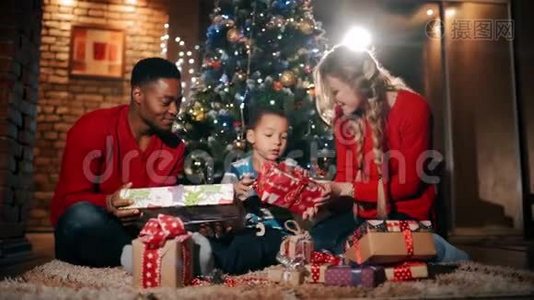 新年礼物中，一个幸福的家庭正坐在圣诞树`附近。 那个男孩正在用红色包装摇动礼物。视频