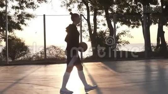 年轻女孩篮球运动员早上在户外的篮球场上散步，同时弹跳球。 侧视图视频