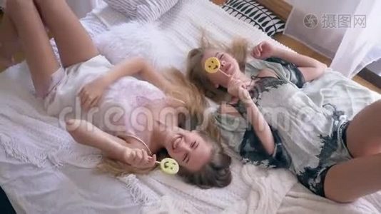 舒适的气氛，穿着丝绸睡衣的姐妹们吃着甜棒棒糖，躺在家里的床上聊天视频