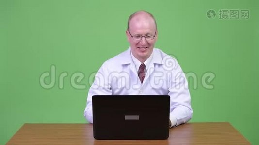 快乐成熟的秃头医生使用笔记本电脑，在木制桌子上得到好消息视频