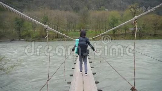 女孩旅行者带着背包在悬索桥上过河，木头渡船过河。 女徒步旅行者视频