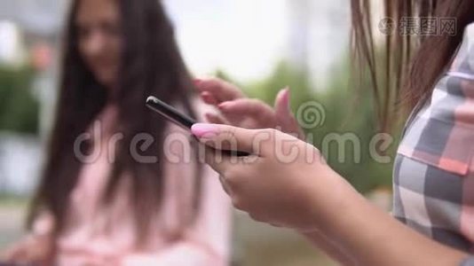 在阳光明媚的天气里，两个年轻女孩坐在喷泉附近的公园里，用智能手机和平板电脑。 慢动作。 高清。 关闭视频