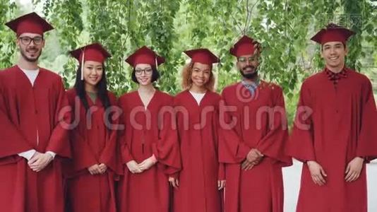 多民族毕业学生站在户外穿着红色长袍和灰泥板、微笑和视频