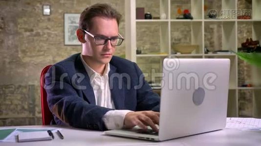 集中的白种人男性坐着看着他的工作笔记本电脑，然后在镜头前放松在砖背景上视频