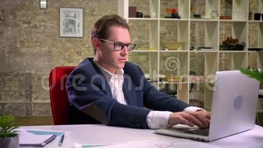 一个疲惫的白种人，在工作场所坐着的时候，双手抱着头，戴着黑色眼镜的笔记本电脑后面视频