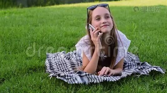 美丽的年轻女孩穿着夏装躺在公园的草地上，使用智能手机。视频