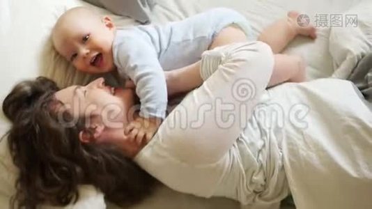 阳光家庭早晨妈妈和宝宝在床上。 一个女人挠痒痒，拥抱她一岁的儿子。 母亲节视频