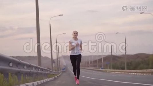 一个穿着白色t恤和轻便运动鞋的运动女孩沿着城外的一条空荡荡的跑道奔跑。 美丽的森林视频