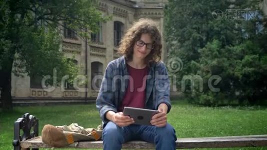 年轻快乐的学生，留着长长的卷发，在平板电脑上打字，坐在大学附近公园的长凳上，面带微笑视频