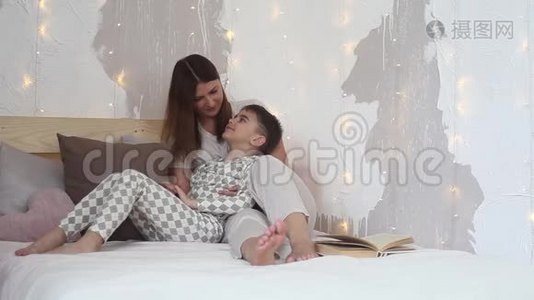 美丽的母亲坐在床上亲吻她的孩子。 孩子躺在母亲`腿上。视频