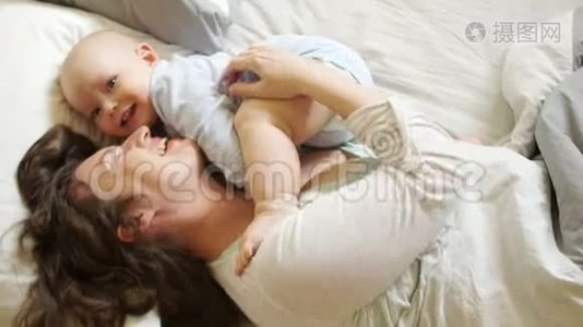 女人挠痒痒，拥抱她那岁的儿子。 阳光家庭早晨妈妈和宝宝在床上。 母亲节视频