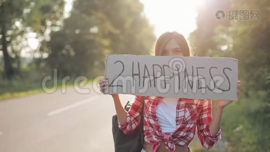 年轻美女搭便车站在路上举着2个幸福的标志。 夏季视频