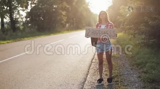 年轻美女搭便车站在路上举着2个幸福的标志。 夏季视频
