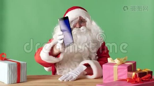 笑容可掬的圣诞老人展示平板电脑，相机里有蓝色屏幕，桌子上有礼物，背景是彩色的。视频